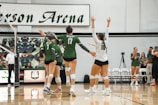 A group of volleyball players in green and white uniforms celebrate on an indoor court. The players are gathered near the net, raising their arms in victory or encouragement. Behind them, a scoreboard and chairs with a logo are visible, along with a few people standing and observing.