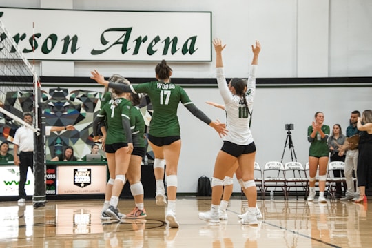 A group of volleyball players in green and white uniforms celebrate on an indoor court. The players are gathered near the net, raising their arms in victory or encouragement. Behind them, a scoreboard and chairs with a logo are visible, along with a few people standing and observing.