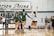 A group of volleyball players in green and white uniforms celebrate on an indoor court. The players are gathered near the net, raising their arms in victory or encouragement. Behind them, a scoreboard and chairs with a logo are visible, along with a few people standing and observing.