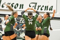 A group of young women wearing green volleyball uniforms celebrating on a volleyball court. The scene appears to be energetic and joyful, with the players showing expressions of excitement and camaraderie. The background includes a geometric-patterned wall and part of the net is visible. The uniforms display numbers and possibly names.