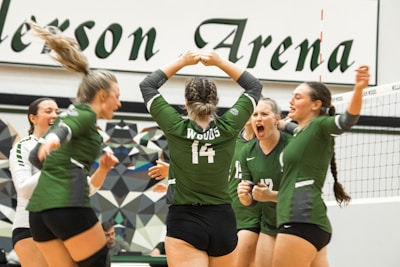 A group of young women wearing green volleyball uniforms celebrating on a volleyball court. The scene appears to be energetic and joyful, with the players showing expressions of excitement and camaraderie. The background includes a geometric-patterned wall and part of the net is visible. The uniforms display numbers and possibly names.
