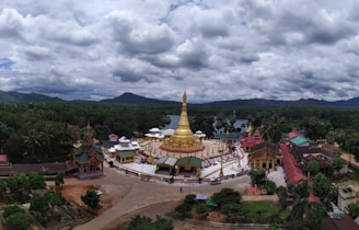 A temple complex with a large golden stupa surrounded by smaller buildings in a lush green landscape. Mountains are visible in the distance under a cloudy sky.