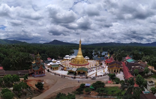A temple complex with a large golden stupa surrounded by smaller buildings in a lush green landscape. Mountains are visible in the distance under a cloudy sky.