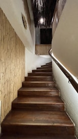 A wooden staircase ascends with a slight curve to the right, flanked by bamboo paneling on one side and a sleek handrail with integrated lighting on the other. The upper portion of the space features rustic, textured elements such as straw or thatch hanging from the ceiling.