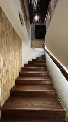 A wooden staircase ascends with a slight curve to the right, flanked by bamboo paneling on one side and a sleek handrail with integrated lighting on the other. The upper portion of the space features rustic, textured elements such as straw or thatch hanging from the ceiling.