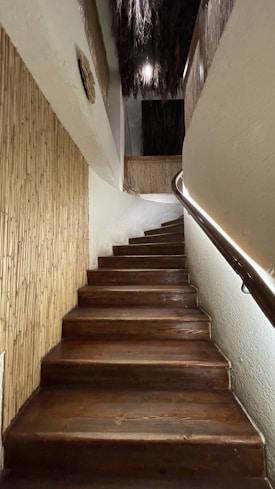 A wooden staircase ascends with a slight curve to the right, flanked by bamboo paneling on one side and a sleek handrail with integrated lighting on the other. The upper portion of the space features rustic, textured elements such as straw or thatch hanging from the ceiling.