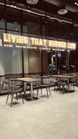 The interior of a modern restaurant with glass walls and wooden tables and chairs. Above the seating area, there is a bright, illuminated sign displaying the phrase 'Living That Burger Life'. The environment appears clean and minimalistic.