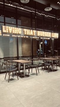 The interior of a modern restaurant with glass walls and wooden tables and chairs. Above the seating area, there is a bright, illuminated sign displaying the phrase 'Living That Burger Life'. The environment appears clean and minimalistic.