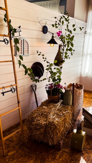A rustic setting featuring a wooden ladder leaning against a white paneled wall. A vintage wall lamp illuminates a climbing plant with leaves and pink flowers. A bale of straw acts as a table, displaying a basket of flowers and metal watering can. A paper bag with text and other containers stand nearby, along with a shovel resting against the wall.