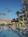 Multiple vibrant, multi-colored buildings with balconies are lined up by a swimming pool. Yellow umbrellas and sun loungers are arranged around the pool. Tall palm trees are scattered around, and clear blue skies are visible in the background.