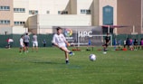 A youth soccer player in a white uniform prepares to make a pass during a practice or game on a green soccer field. Other players and coaches are present in the background, along with a large building and a banner with logos. There is sunlight suggesting a bright, clear day.
