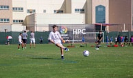 A youth soccer player in a white uniform prepares to make a pass during a practice or game on a green soccer field. Other players and coaches are present in the background, along with a large building and a banner with logos. There is sunlight suggesting a bright, clear day.
