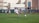 A youth soccer player in a white uniform prepares to make a pass during a practice or game on a green soccer field. Other players and coaches are present in the background, along with a large building and a banner with logos. There is sunlight suggesting a bright, clear day.