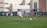 A youth soccer player in a white uniform prepares to make a pass during a practice or game on a green soccer field. Other players and coaches are present in the background, along with a large building and a banner with logos. There is sunlight suggesting a bright, clear day.