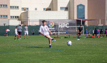 A youth soccer player in a white uniform prepares to make a pass during a practice or game on a green soccer field. Other players and coaches are present in the background, along with a large building and a banner with logos. There is sunlight suggesting a bright, clear day.