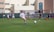A youth soccer player in a white uniform prepares to make a pass during a practice or game on a green soccer field. Other players and coaches are present in the background, along with a large building and a banner with logos. There is sunlight suggesting a bright, clear day.