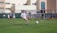 A youth soccer player in a white uniform prepares to make a pass during a practice or game on a green soccer field. Other players and coaches are present in the background, along with a large building and a banner with logos. There is sunlight suggesting a bright, clear day.
