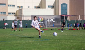 A youth soccer player in a white uniform prepares to make a pass during a practice or game on a green soccer field. Other players and coaches are present in the background, along with a large building and a banner with logos. There is sunlight suggesting a bright, clear day.