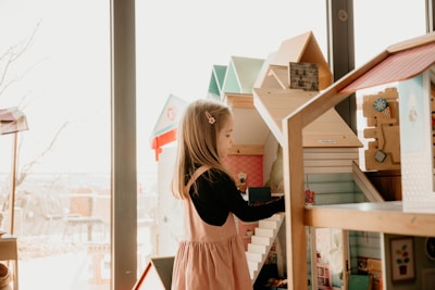 A young girl with long hair, wearing a pink dress and a black shirt, is playing with a detailed wooden dollhouse. The dollhouse features several rooms and stairs, with various small furniture pieces. The background includes large windows letting in natural light, and other play structures are partially visible.