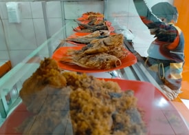 A row of red plates, each holding a fried fish, is displayed in a glass cabinet. The setting seems to be a food stall or market. There is a person wearing warm clothing and a hat visible behind the counter.