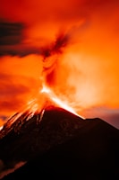 A towering volcano spews bright lava against an intense orange sky, with ash and smoke billowing dramatically upward. The fiery glow highlights the rugged contours of the volcanic peak, creating a striking contrast with the darkened foreground.