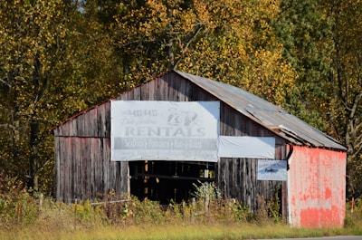 A weathered, rustic barn with a peeling wooden exterior sits amongst a backdrop of dense autumn foliage. The barn has an old sign that advertises rentals for watercraft, suggesting it's located near a body of water. The roof is corrugated and shows signs of age, with rust and wear. Tall grass and wild plants surround the structure, adding to the rural, untamed feel of the scene.