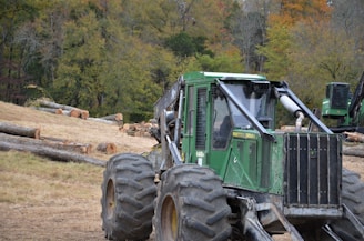 A heavy-duty green forestry machine is positioned on uneven terrain with large tires and a strong metallic body. It's surrounded by a logging area with numerous cut tree trunks scattered on the ground. In the background, there is a dense forest with trees displaying autumn colors.