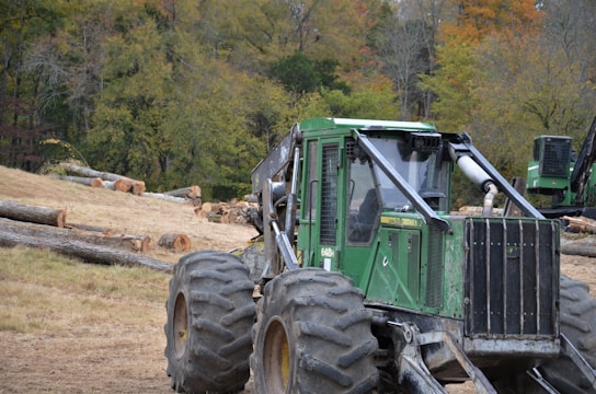 A heavy-duty green forestry machine is positioned on uneven terrain with large tires and a strong metallic body. It's surrounded by a logging area with numerous cut tree trunks scattered on the ground. In the background, there is a dense forest with trees displaying autumn colors.