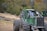 A heavy-duty green forestry machine is positioned on uneven terrain with large tires and a strong metallic body. It's surrounded by a logging area with numerous cut tree trunks scattered on the ground. In the background, there is a dense forest with trees displaying autumn colors.