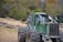 A heavy-duty green forestry machine is positioned on uneven terrain with large tires and a strong metallic body. It's surrounded by a logging area with numerous cut tree trunks scattered on the ground. In the background, there is a dense forest with trees displaying autumn colors.