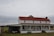 A rustic, two-story farmhouse with a red metal roof and white wooden siding. The house features a long front porch with several chairs and a small table. There is a covered area for a vehicle beside it, and a green field stretches out in the background under an overcast sky.