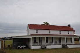A rustic, two-story farmhouse with a red metal roof and white wooden siding. The house features a long front porch with several chairs and a small table. There is a covered area for a vehicle beside it, and a green field stretches out in the background under an overcast sky.