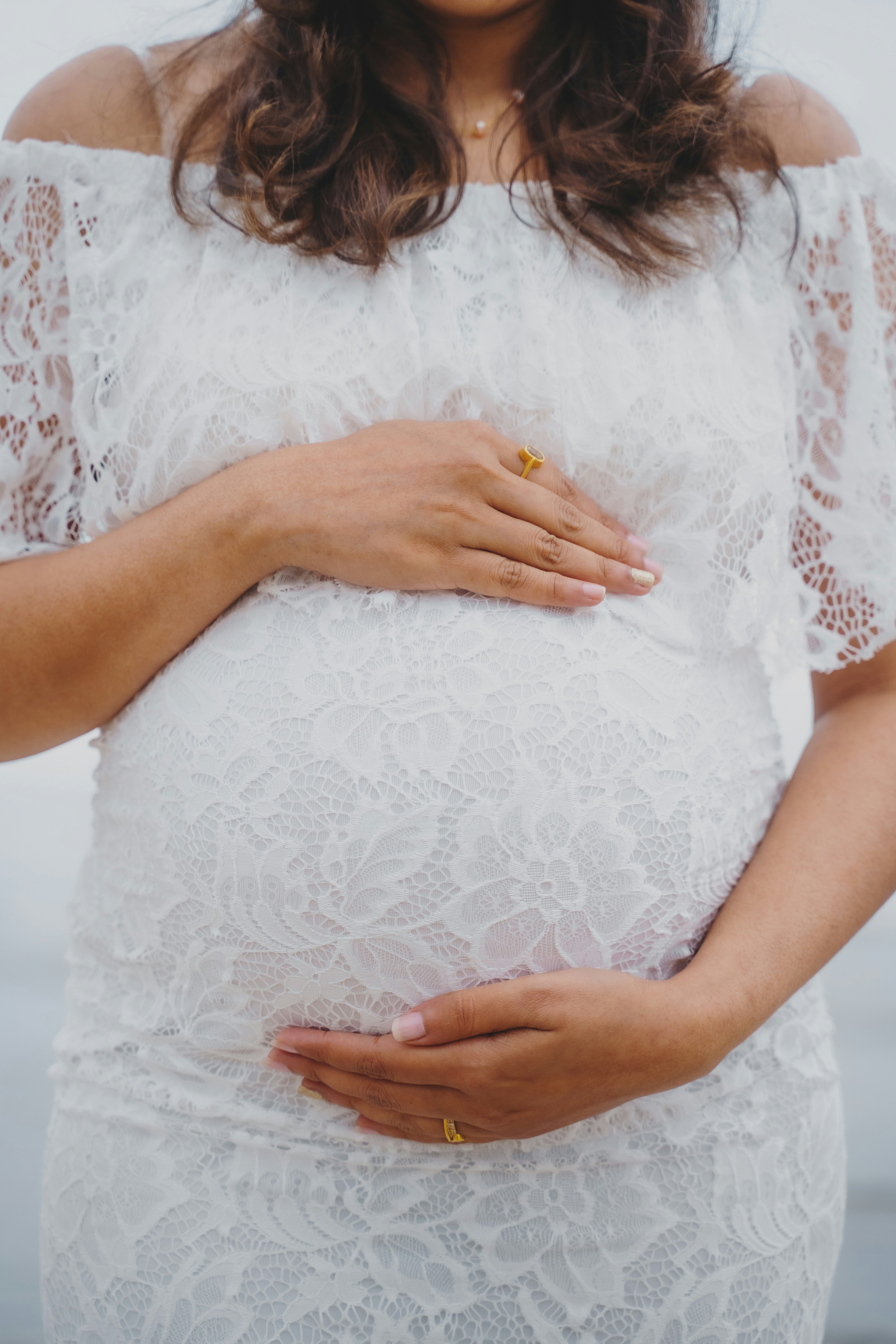 Mother in a white gown holding her baby bump standing next to a beautiful beach.