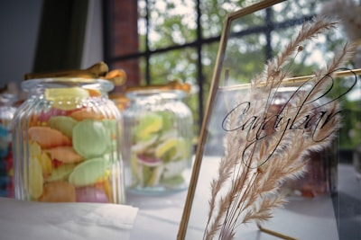 Jars filled with colorful candies are displayed on a table, accompanied by a gold-framed glass sign with the word 'candybar' written on it. The background shows a window with greenery visible outside, and decorative dried grasses placed behind the sign.