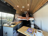 An interior view of a van converted into a cozy living space. A woman is sitting on a bed with orange print bedding, holding a mug. The kitchen area on the left includes a sink, a faucet, and various kitchen items. A wooden ceiling adds warmth to the space, and a magazine on the table features a cover about Everest.