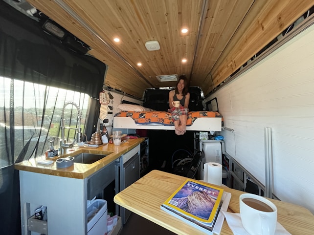 An interior view of a van converted into a cozy living space. A woman is sitting on a bed with orange print bedding, holding a mug. The kitchen area on the left includes a sink, a faucet, and various kitchen items. A wooden ceiling adds warmth to the space, and a magazine on the table features a cover about Everest.
