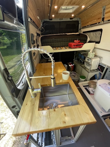A van interior converted into a living space featuring a wooden countertop with a built-in stainless steel sink. There is a flexible metal faucet and a white cup placed on the counter. In the background, an elevated bed platform is visible with a mattress and a red toolbox. Shelving and storage units are mounted along the walls, providing ample organization.