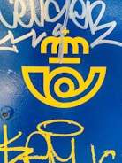 Bright yellow logo with a postal emblem design and a crown above, set against a vivid blue background. White and yellow graffiti marks are present over the surface.