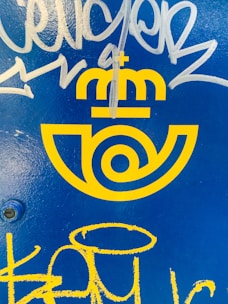 Bright yellow logo with a postal emblem design and a crown above, set against a vivid blue background. White and yellow graffiti marks are present over the surface.