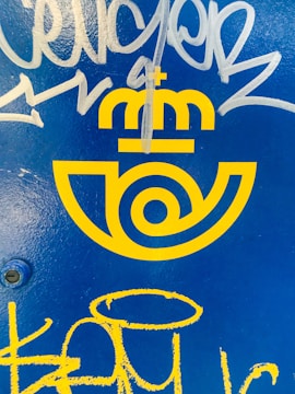 Bright yellow logo with a postal emblem design and a crown above, set against a vivid blue background. White and yellow graffiti marks are present over the surface.