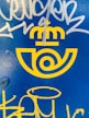 Bright yellow logo with a postal emblem design and a crown above, set against a vivid blue background. White and yellow graffiti marks are present over the surface.