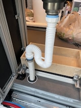 A flexible white hose connects to a metal sink drain. It is looped in an S-shape secured with fastening rings. The setup is placed within a metallic and wooden frame, which appears to be part of cabinetry or a storage unit. In the background, there are plastic bags and other household items.