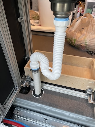 A flexible white hose connects to a metal sink drain. It is looped in an S-shape secured with fastening rings. The setup is placed within a metallic and wooden frame, which appears to be part of cabinetry or a storage unit. In the background, there are plastic bags and other household items.