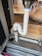 A flexible white hose connects to a metal sink drain. It is looped in an S-shape secured with fastening rings. The setup is placed within a metallic and wooden frame, which appears to be part of cabinetry or a storage unit. In the background, there are plastic bags and other household items.