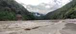 A wide river flows between two lush, green mountainous landscapes. The water appears to be moving with some force, creating small waves across the surface. The mountains are densely covered with trees and vegetation. Clouds hang low in the sky, partially obscuring the mountain peaks.