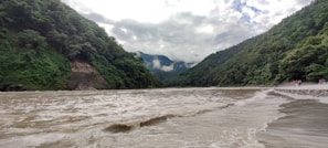 A wide river flows between two lush, green mountainous landscapes. The water appears to be moving with some force, creating small waves across the surface. The mountains are densely covered with trees and vegetation. Clouds hang low in the sky, partially obscuring the mountain peaks.