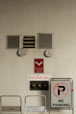 A beige tiled wall contains various fire safety and parking signs. Two sprinkler fire alarm circular signs are mounted on the wall above red signs indicating a fire department connection. There is a prominent 'NO PARKING' sign to the right side, and two wall vents and a central air vent are also visible.