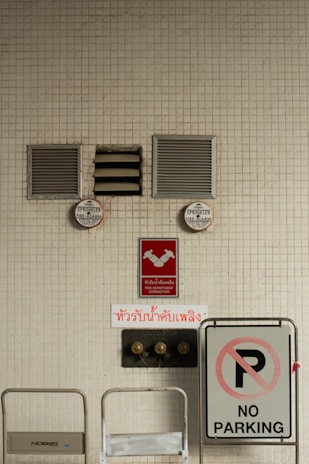 A beige tiled wall contains various fire safety and parking signs. Two sprinkler fire alarm circular signs are mounted on the wall above red signs indicating a fire department connection. There is a prominent 'NO PARKING' sign to the right side, and two wall vents and a central air vent are also visible.