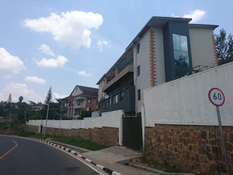A modern multi-story building with glass panelling stands beside a curved road. The road is bordered by a stone and white wall, with a speed limit sign indicating 60. In the background, another brick building with a triangular roof is partially visible, surrounded by green trees. The sky is bright with scattered clouds.