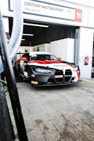 A racing car with a black, red, and white color scheme is positioned in a motorsport garage under a sign that reads 'Century Motorsport BMW M4 GT3'. The car has various brand stickers, including Sonax and Pirelli, and is partially outside the garage ready for a race.